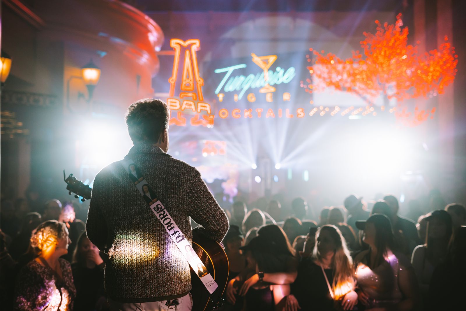 Musician playing guitar on stage facing a lively crowd under neon signs at Turquoise Tiger Bar and Cocktails.