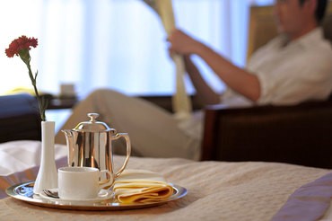 Room service dining tray with Coffee and pastry being enjoyed while a man reads the paper in his hotel room