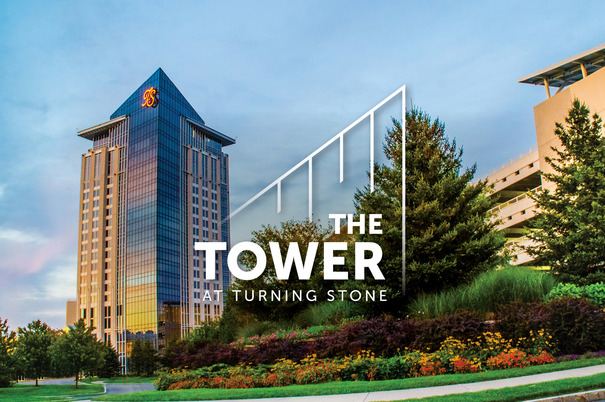 Trees and flowers line the approach to The Tower at Turning Stone. The setting sun illuminates the glass on The Tower.