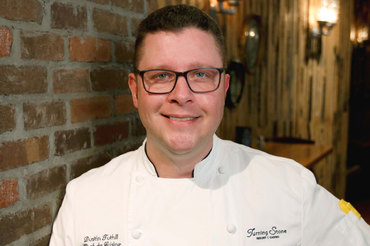 Head and shoulder portrait of Chef Dustin Tuthill wearing chef jacket in front of red brick wall