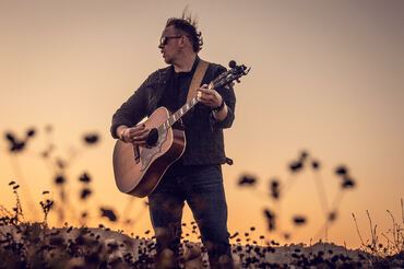 Alec MacGillivray playing guitar in a field of flowers at sunset
